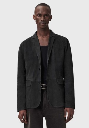 Zwarte suède blazer met twee voorzakken, inkeping revers en sluiting met één knoop. Draagt over een zwart T-shirt en gecombineerd met donkere jeans.