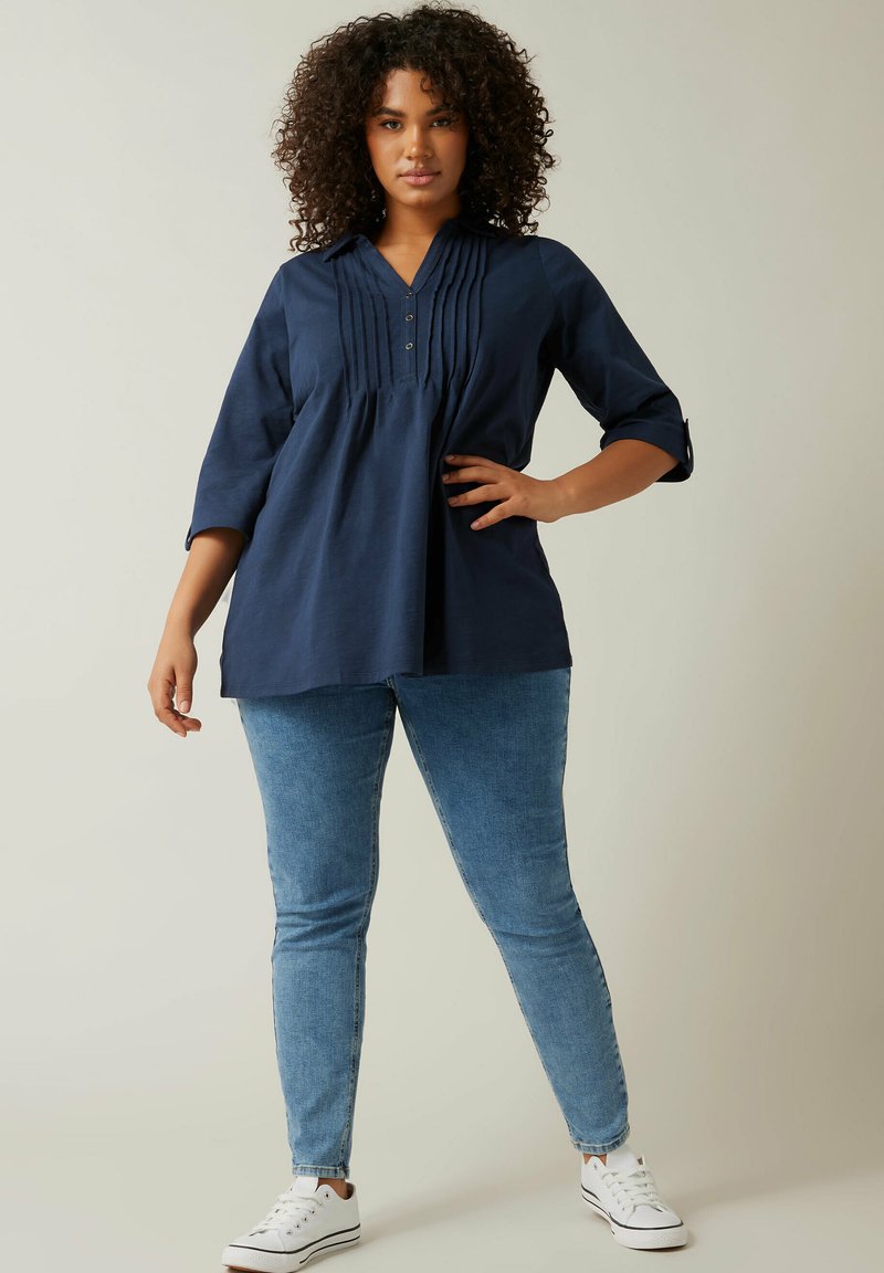 Top blu navy a tre quarti con pences al collo, abbottonatura e vestibilità comoda, abbinato a jeans azzurri chiari e sneaker bianche.