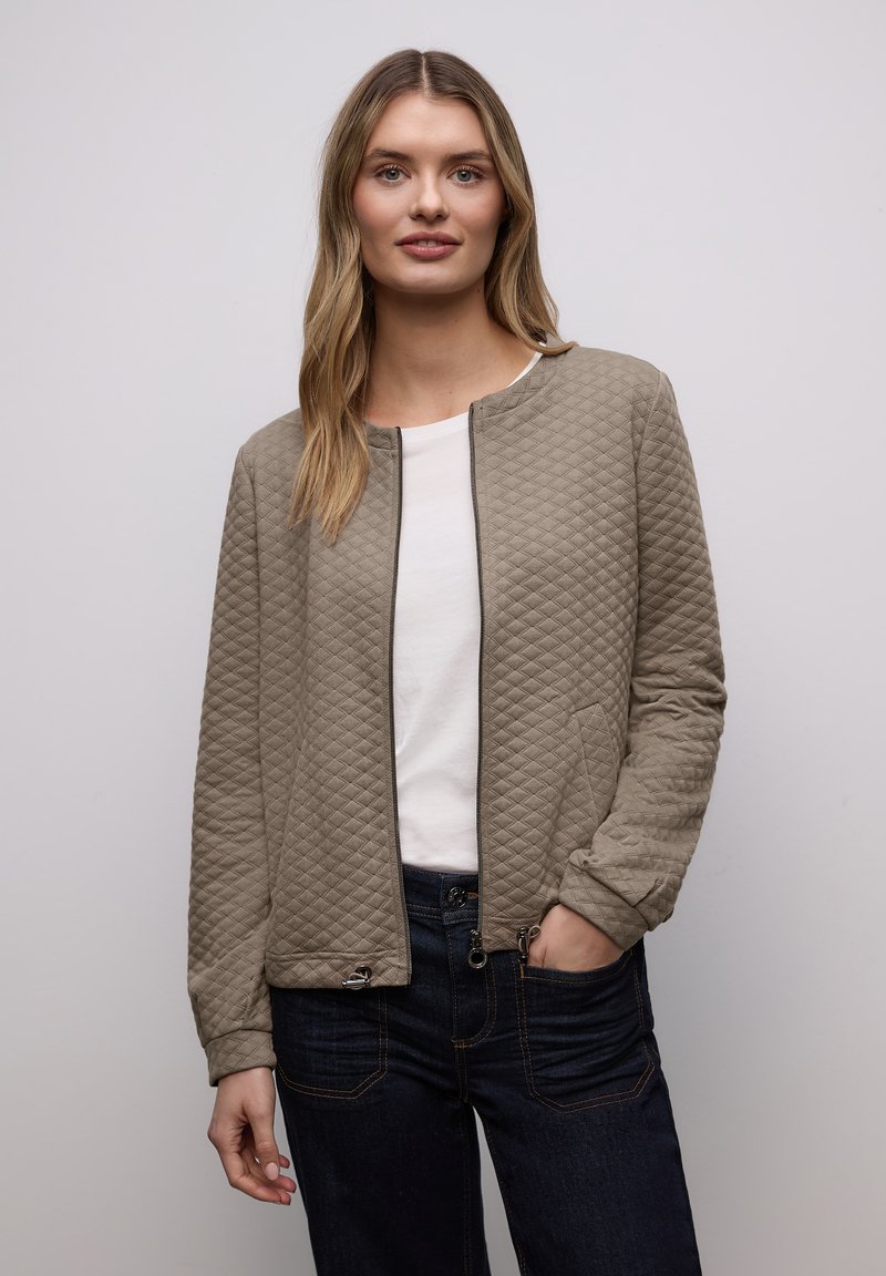 Gesteppte beige Jacke mit rundem Ausschnitt, Reißverschluss vorne und dezenter Struktur. Getragen über einem weißen Oberteil mit dunkelblauen Jeans.
