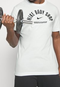 Biała koszulka sportowa z czarnym napisem "Nike Body Shop" i szachownicowym detalem, trzymana w ręce chwycającej czarny hantel.