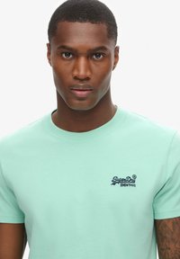 Mintgrøn bomulds t-shirt med korte ærmer og ribbet rund halsudskæring. "SuperDry" logoet er broderet på venstre bryst i marineblå.