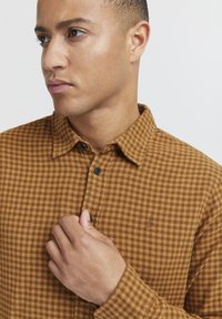 Camisa de cuadros marrón de botones, hecha de un tejido texturizado. Presenta un cuello, botones negros y un pequeño logotipo bordado en el pecho.