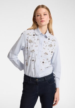 Camicia azzurro chiaro con maniche lunghe, decorata con ricami floreali argentati sul davanti, abbinata a jeans blu scuro.
