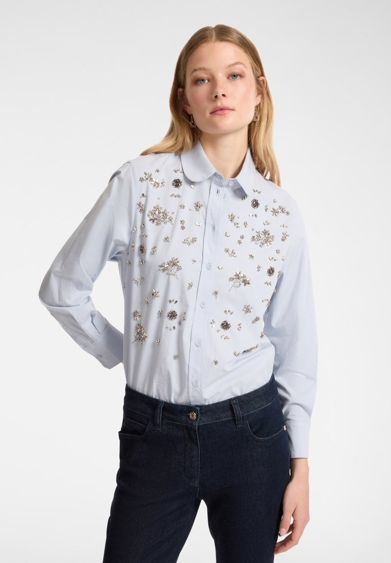 Camicia azzurro chiaro con maniche lunghe, decorata con ricami floreali argentati sul davanti, abbinata a jeans blu scuro.