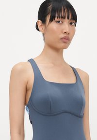 Maillot de bain une pièce bleu avec des bretelles larges, un décolleté carré et des accents cousus, présentant une texture lisse et ajustée.