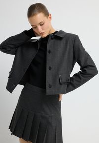 Czarna krótka kurtka z błyszczącym czarnym kołnierzem, z zapięciem na guziki i akcentowanymi kieszeniami; zestawiona z dopasowaną czarną spódnicą plisowaną z prążkami.