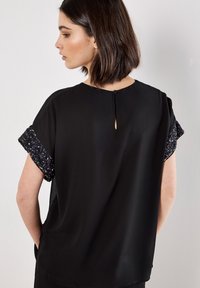 Schwarzes kurzärmliges Oberteil mit Paillettenakzenten an den Ärmeln, mit einer Knopfverschluss hinten und glatter Textur.