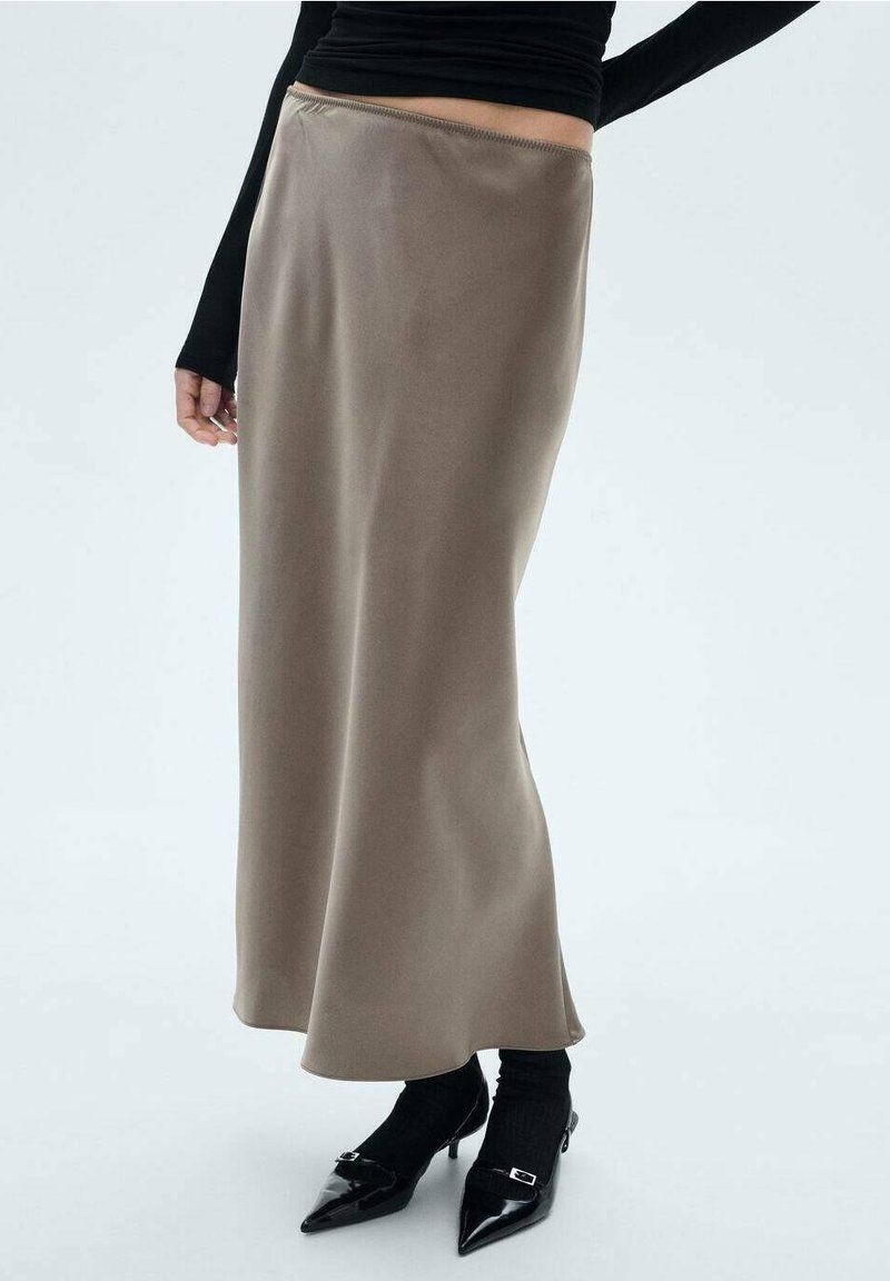 Bruine satijnen maxi-jurk met een soepele textuur, hoge taille en een vloeiende silhoutte, gecombineerd met zwarte schoenen en aangesloten lange mouwen.