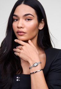 Montre argentée avec un cadran carré et un bracelet à maillons, accompagnée d'un bracelet noir fin avec des accents circulaires. Modèle portant un haut noir minimaliste.