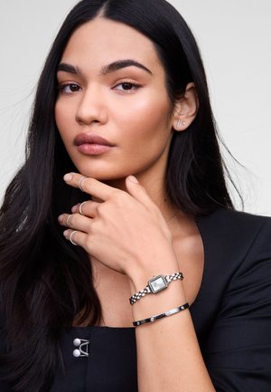 Montre argentée avec un cadran carré et un bracelet à maillons, accompagnée d'un bracelet noir fin avec des accents circulaires. Modèle portant un haut noir minimaliste.