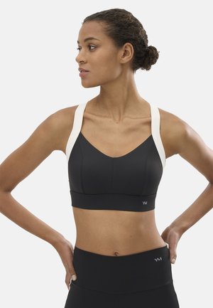 Mujer con sujetador deportivo negro con tirantes blancos y leggins de talle alto a juego, con las manos apoyadas en las caderas y mirando hacia un lado.