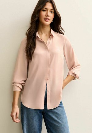 Jasnoróżowa jedwabna koszula na guziki z długimi rękawami, o luźnym kroju i podwiniętymi mankietami, zestawiona z niebieskimi dżinsami.