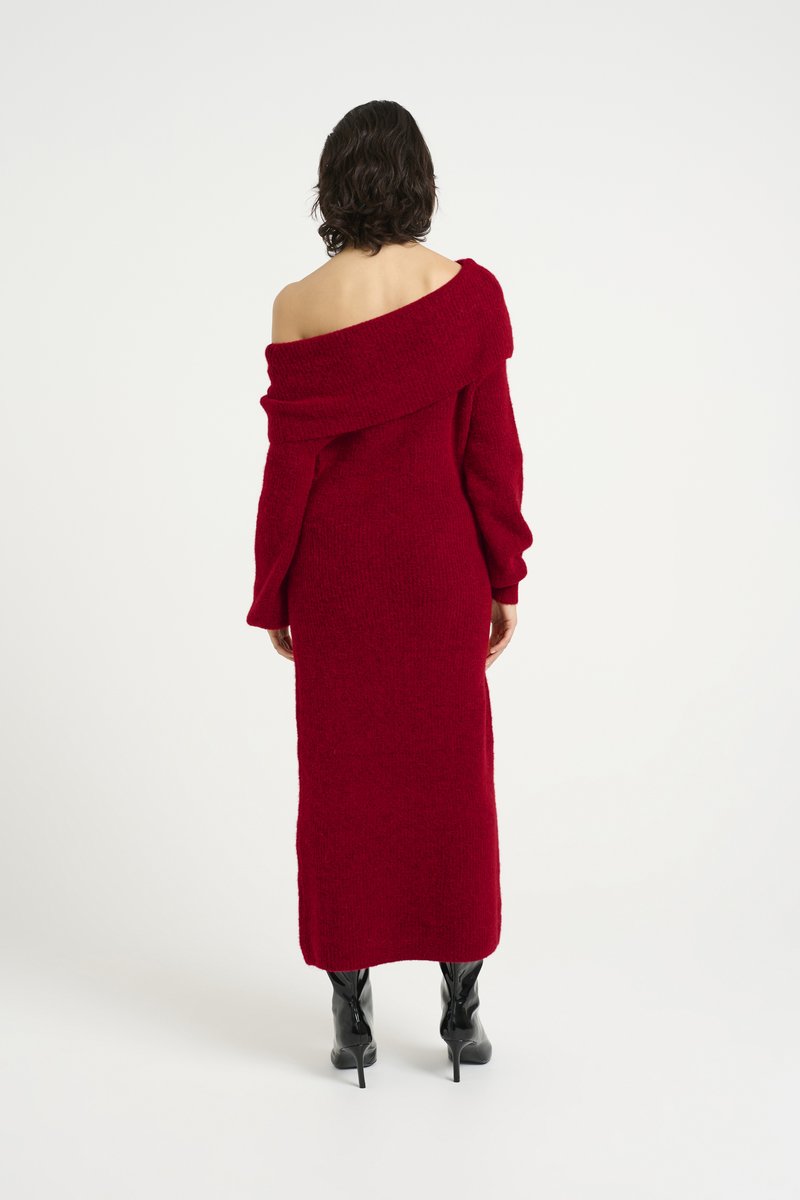 Langärmliges, rotes Strickkleid mit Off-Shoulder-Ausschnitt, das aus einem strukturierten Stoff gefertigt ist. Die Passform ist figurbetont und fällt bis zur Mitte der Wade. Schwarze, hochhackige Stiefel.