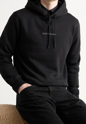 Mężczyzna w czarnej bluzie Calvin Klein Jeans i czarnych spodniach, z jedną ręką w kieszeni, siedzący na teksturowanej, brązowej powierzchni.