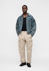 Άνδρας με μπλε τζιν μπουφάν, μαύρο πουκάμισο, μπεζ παντελόνι καμπαρντίνας και μαύρα αθλητικά παπούτσια, στέκεται με τα χέρια στις τσέπες μπροστά σε λευκό φόντο.