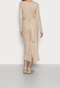 Frau in einem beigen Midi-Kleid mit langen Ärmeln und einem gebundenen Gürtel in der Taille sowie beigen Sneakers, von hinten gezeigt.