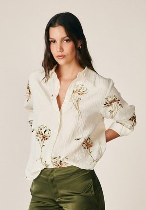 Mujer con cabello largo y oscuro lleva una blusa crema con bordado floral y pantalones verde oliva, de pie con una mano en la cadera.