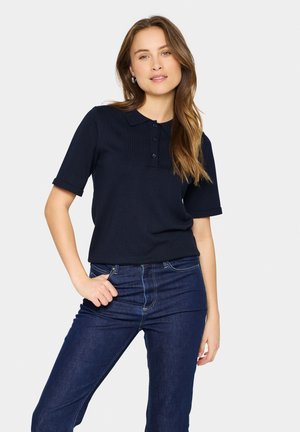 Navyblaues Polohemd mit kurzen Ärmeln, einem knöpfbaren Ausschnitt und geripptem Material, kombiniert mit dunklen Jeans im klassischen Schnitt.