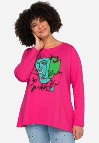 Camiseta de manga larga color rosa con un diseño gráfico de rostros abstractos en verde y azul, y el texto "Dormir para soñar" en negro.