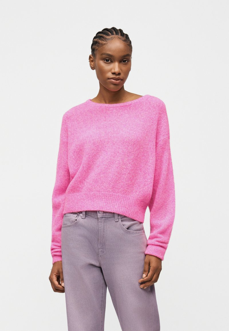 Γυναίκα με κοτσίδες, που φοράει φωτεινό ροζ χνουδωτό πουλόβερ και ανοιχτό μωβ τζιν ψηλόμεσο, στέκεται μπροστά σε απλό λευκό φόντο.