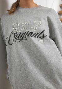 Hallhalli hallituskamas, millel on bold-tähtedega "SELF Originals", pehme tekstuur ja mugav lõige. Aastal "2023" väiksemas kirjas.