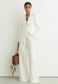 Mujer con el cabello trenzado, vestida con un traje blanco a medida y pantalones de pierna ancha, sosteniendo un bolso marrón con una bufanda a rayas.
