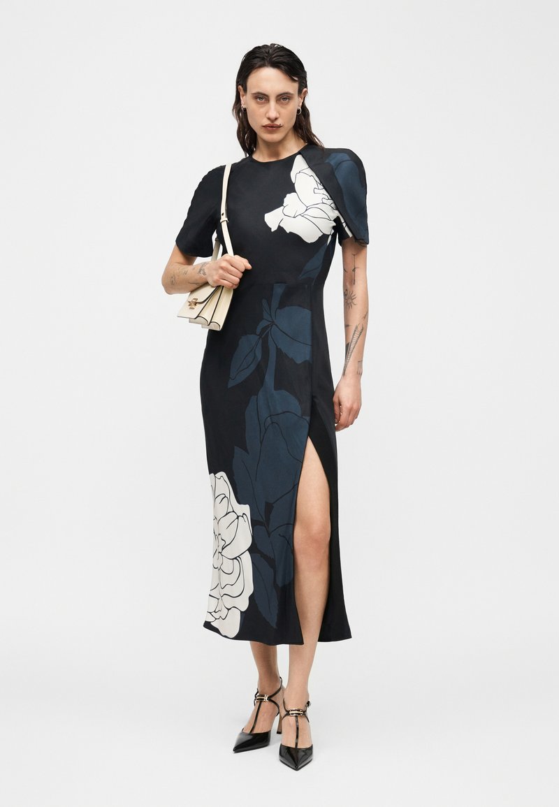 Femme portant une robe midi noire avec de grands motifs floraux blancs et bleu foncé, épaules bouffantes, fente à la cuisse, talons noirs, et tenant un petit sac beige.