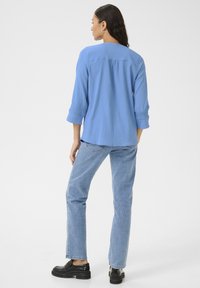 Chemise texturée bleu clair, avec une coupe décontractée, manches 3/4 et ourlet arrondi, accompagnée d'un jean en denim clair et de chaussures noires.