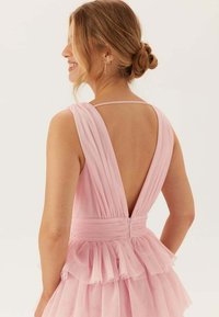 Różowa sukienka z głębokim dekoltem w kształcie litery V na plecach, z plisowanym materiałem i warstwowym falbankowym dołem. Cienkie ramiączka i zamek błyskawiczny z tyłu.