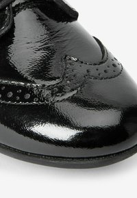 Schwarzer Lacklederschuh mit geschwungenem Design, durchbrochenen Details und dekorativen Nähten entlang der Kanten. Glatte, glänzende Textur.