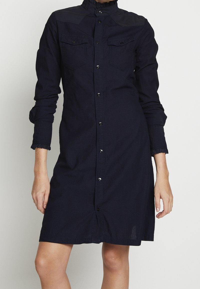 Robe bleu marine à boutons avec des manches longues, col volanté, deux poches poitrine et poignets effilochés, fabriquée en tissu texturé.