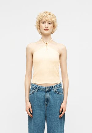 Młoda osoba z krótkimi kręconymi blond włosami, ubrana w jasny top na ramiączkach i wysokim stanem niebieskie dżinsy, stoi na tle jednolitego białego tła.