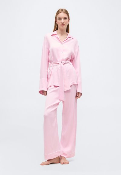 Γυναίκα που στέκεται ξυπόλητη φορώντας χαλαρό ροζ σατέν πουκάμισο με μακριά μανίκια και ζώνη, καθώς και παντελόνι με φαρδιά πόδια σε αντίστοιχο χρώμα, σε απλό φόντο.