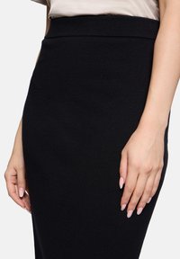 Femme portant une jupe noire ajustée taille haute et un haut beige, les mains reposant doucement sur les côtés de la jupe.