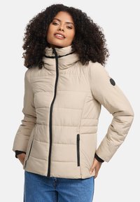 Vrouw in een beige gewatteerde jas met zwarte ritsdetails en een spijkerbroek, staand met handen in de zakken.