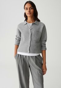Femme portant un cardigan gris à côtes avec des boutons et une grande poche avant, une chemise blanche en dessous, et un pantalon gris plissé avec les mains dans les poches.
