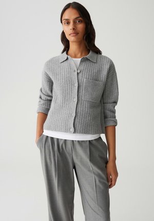 Femme portant un cardigan gris à côtes avec des boutons et une grande poche avant, une chemise blanche en dessous, et un pantalon gris plissé avec les mains dans les poches.