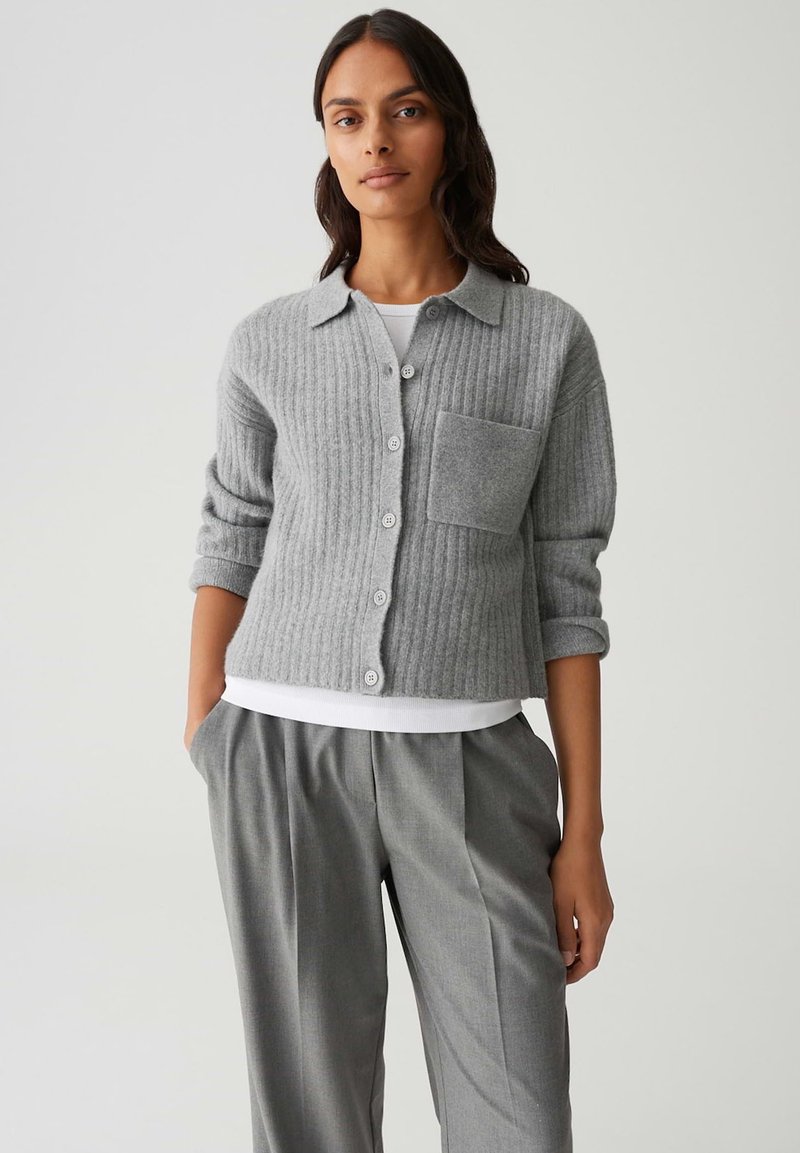 Femme portant un cardigan gris à côtes avec des boutons et une grande poche avant, une chemise blanche en dessous, et un pantalon gris plissé avec les mains dans les poches.