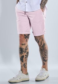 Light pink cotton shorts with a relaxed fit and rolled hems, paired with white and green sneakers. Legs feature detailed tattoos.