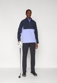 Pulover pentru bărbați, de culoare bleumarin și mov deschis, cu fermoar pe un sfert, mâneci lungi și material solid; purtat cu pantaloni închiși la culoare și un club de golf.