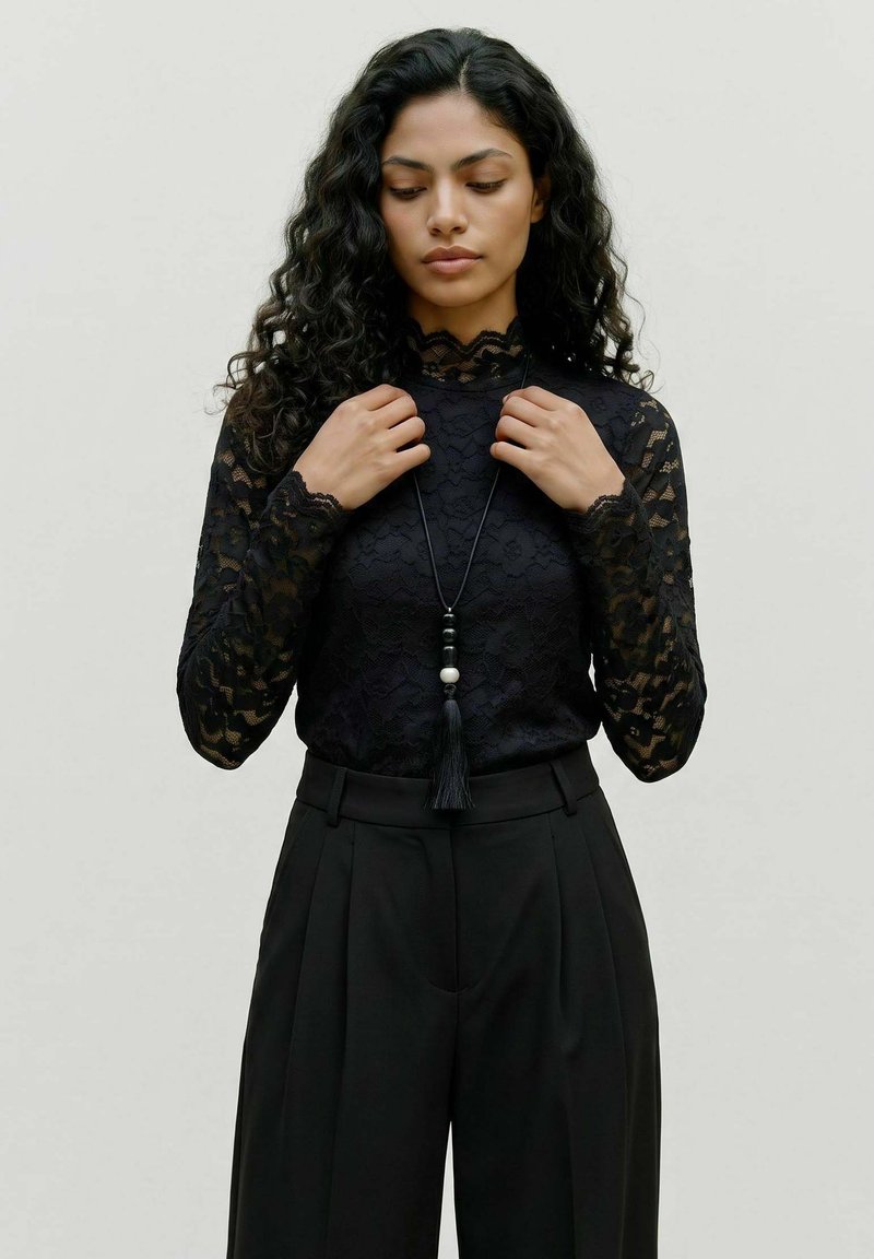 Femme aux longs cheveux bouclés portant un haut en dentelle noire à manches longues et un pantalon noir taille haute, ajustant un collier avec un pendentif à pompon.