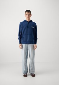 Felpa con cappuccio blu navy, dotata di tasca frontale e cordini, abbinata a jeans larghi e chiari e scarpe marroni scure.