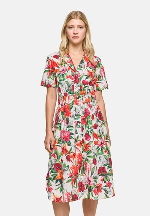 Woman standing wearing a knee-length, short-sleeve dress with a colorful floral pattern, looking forward against a plain light background.
