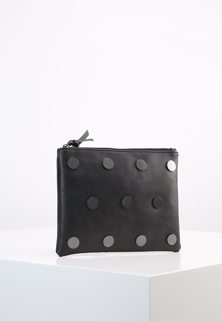 Pochette rectangulaire noire avec des clous métalliques ronds disposés en trois rangées et une fermeture éclair sur le dessus, posée sur une surface blanche.