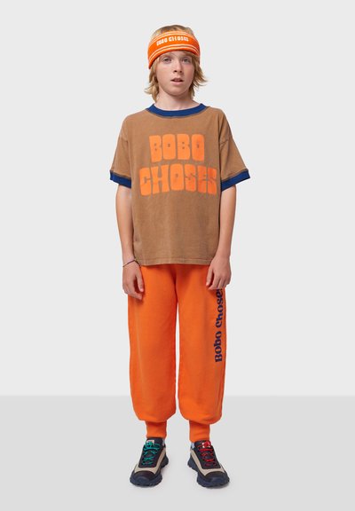 Braunes, übergroßes T-Shirt mit orangefarbenem "BOBO CHOSES"-Text, kombiniert mit orangefarbenen Jogginghosen, die ein seitliches Logo in Blau tragen. Schwarze Sneakers mit bunten Schnürsenkeln.