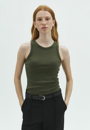 Young person with long red hair wearing a sleeveless green top and black pants, standing with hands in pockets against a plain background.