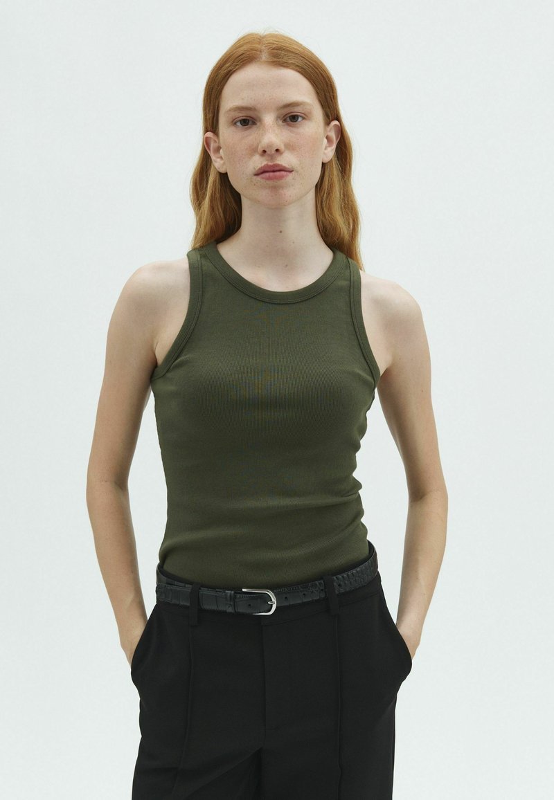 Jeune personne aux longs cheveux roux portant un haut vert sans manches et un pantalon noir, debout les mains dans les poches devant un fond uni.