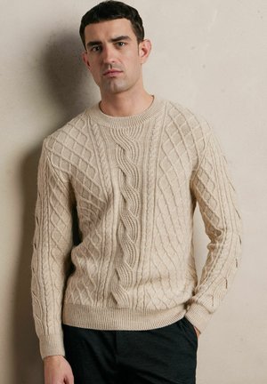 Beżowy sweter dzianinowy z teksturowanym wzorem w warkocze, z okrągłym dekoltem oraz ściągaczami przy mankietach i u dołu. Noszony na ciemnych spodniach.