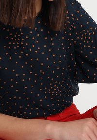 Marinebluse mit braunen Pünktchen, kurzen Ärmeln und rundem Halsauschnitt. Glatte Textur mit lockerer Passform, kombiniert mit roten Hosen.