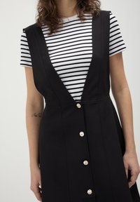 Robe noire sans manches avec un décolleté en V profond, ornée de boutons argentés sur le devant, portée par-dessus une chemise rayée noire et blanche.